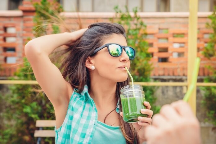 Woman with sunglasses drinking green vegetable smoothie outdoors
